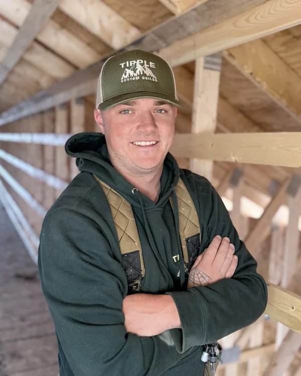 Josh Reidl, carpenter, arms crossed in a framed attic in his company cap and tool belt.