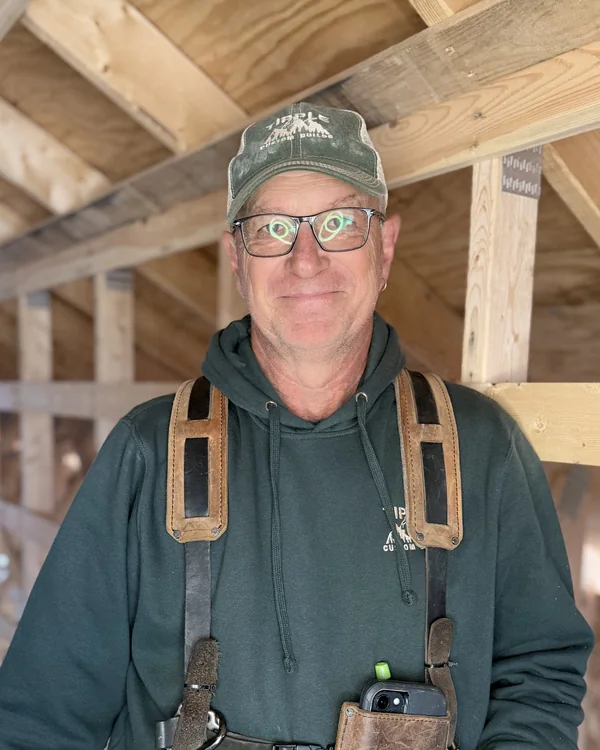 Scott Tipple, carpenter, smiling in a framed attic in his company cap and leather tool belt.