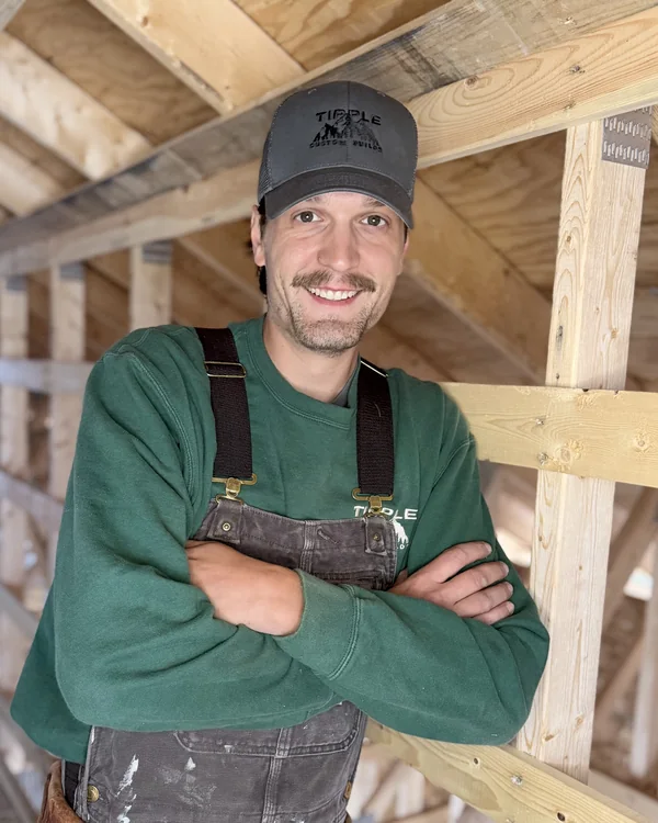 Skyler Kato, arms crossed against the framing in a company cap and paint-marked overalls.