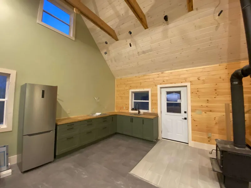 Green shaker cabinets and a wood counter against pine-boarded walls, wood stove alongside.