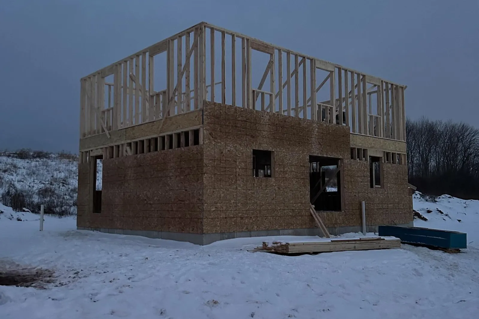 A house framed to the second-floor deck on a snowy lot, sheathed and waiting on trusses.