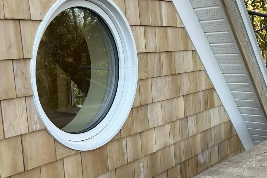 Cedar shingle siding coursed around a round porthole window under a raking roof line.
